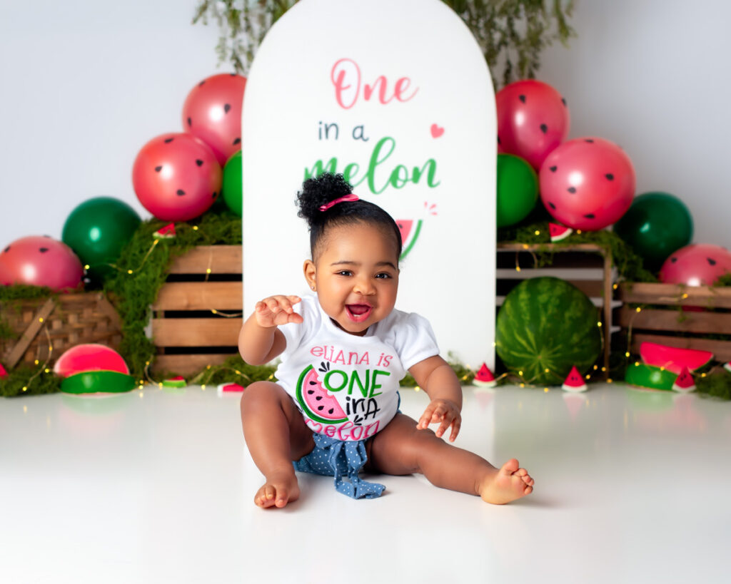 Smiling baby sitting in a watermelon-themed cake smash set with pink and green balloon decorations