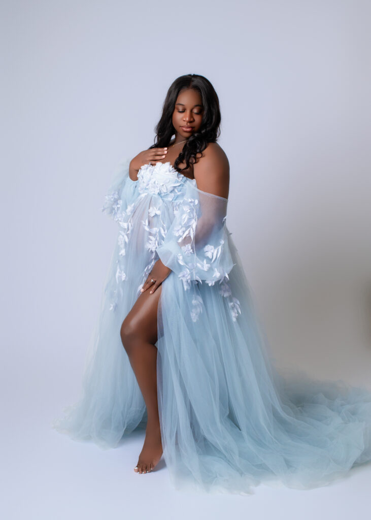 Pregnant woman in flowing blue gown, looking down at her shoulder with a serene expression during a studio maternity session.