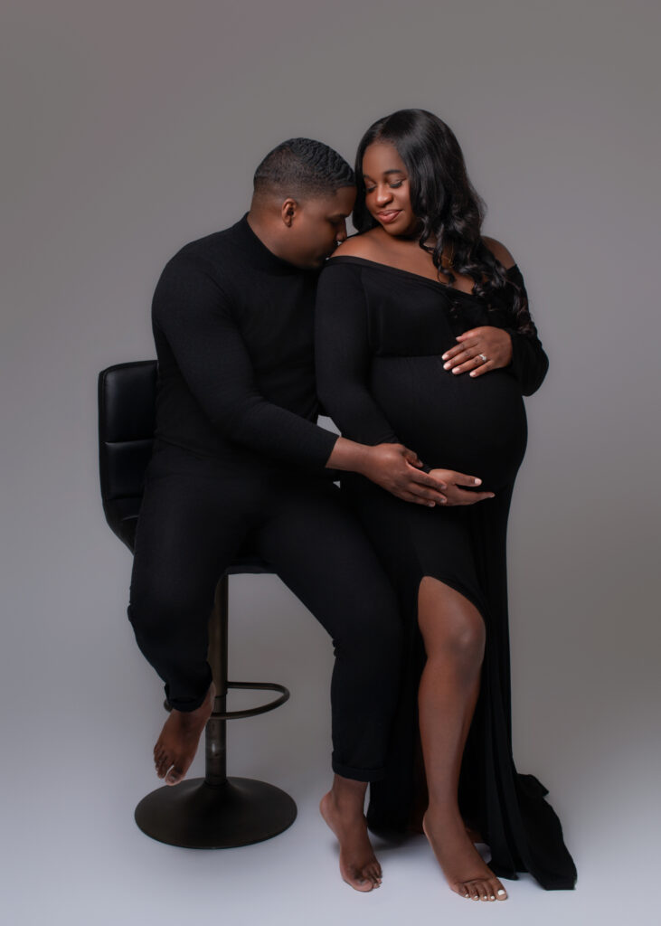 Couple in black formal outfits sitting together with foreheads touching during maternity session.