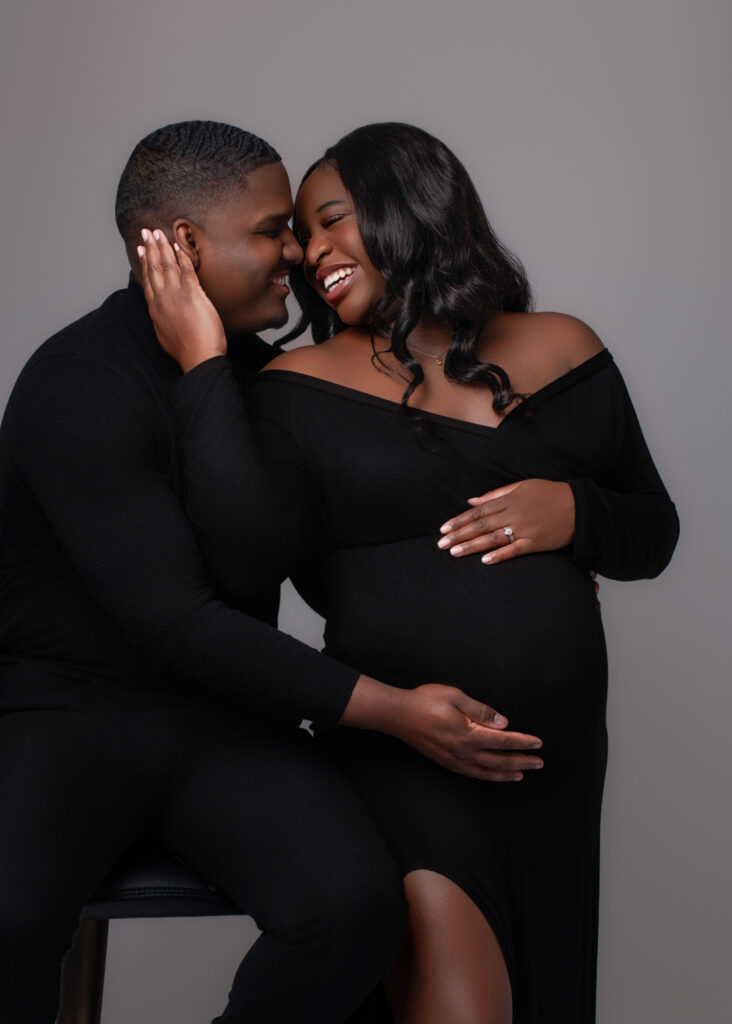 Pregnant woman in black gown sits beside her husband, smiling at each other with joyful connection during their maternity session.