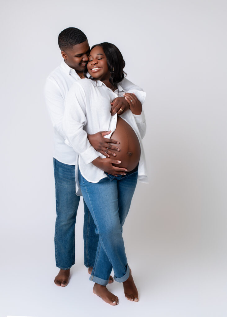 Maternity photos with husband in casual white shirts and jeans, smiling and embracing during a studio session