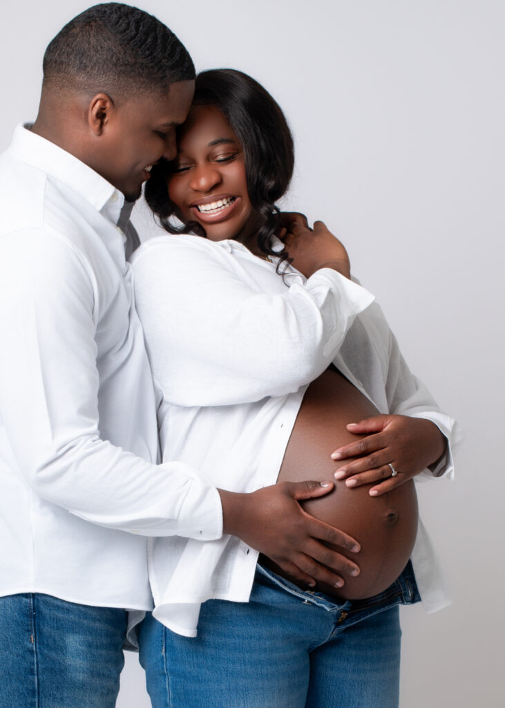 Maternity photo with husband standing close and resting his hand on her belly while she looks down, eyes closed in a tender moment.
