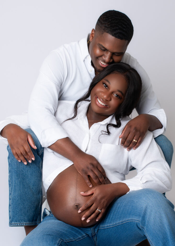 Maternity photo of husband sitting behind his wife, arms wrapped around her belly in a cozy studio portrait.