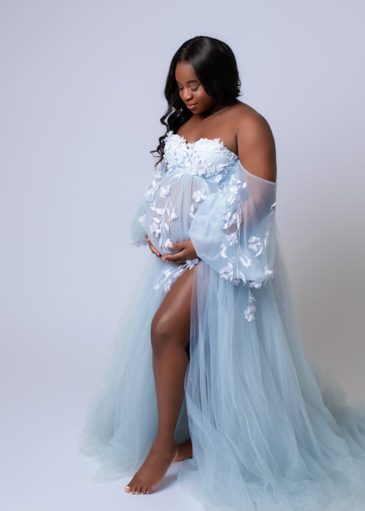 Pregnant woman in a pale blue floral tulle gown standing barefoot in a soft studio setting, gently holding her belly.