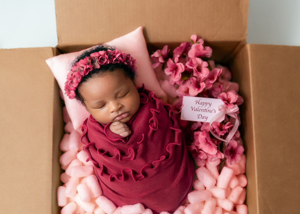 baby wrapped in fuchsia ruffles with a flower halo laying in a box full of pink peanuts and flowers with a Happy Valentine's day tag.