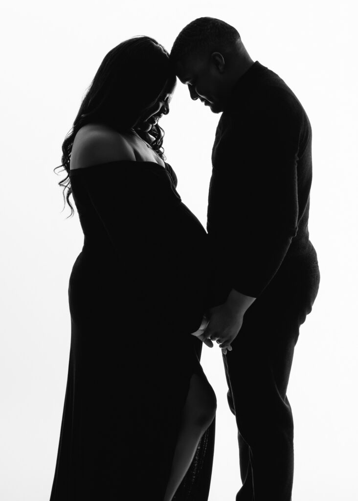 Silhouette of expecting couple standing forehead to forehead, holding hands in a quiet moment of connection.