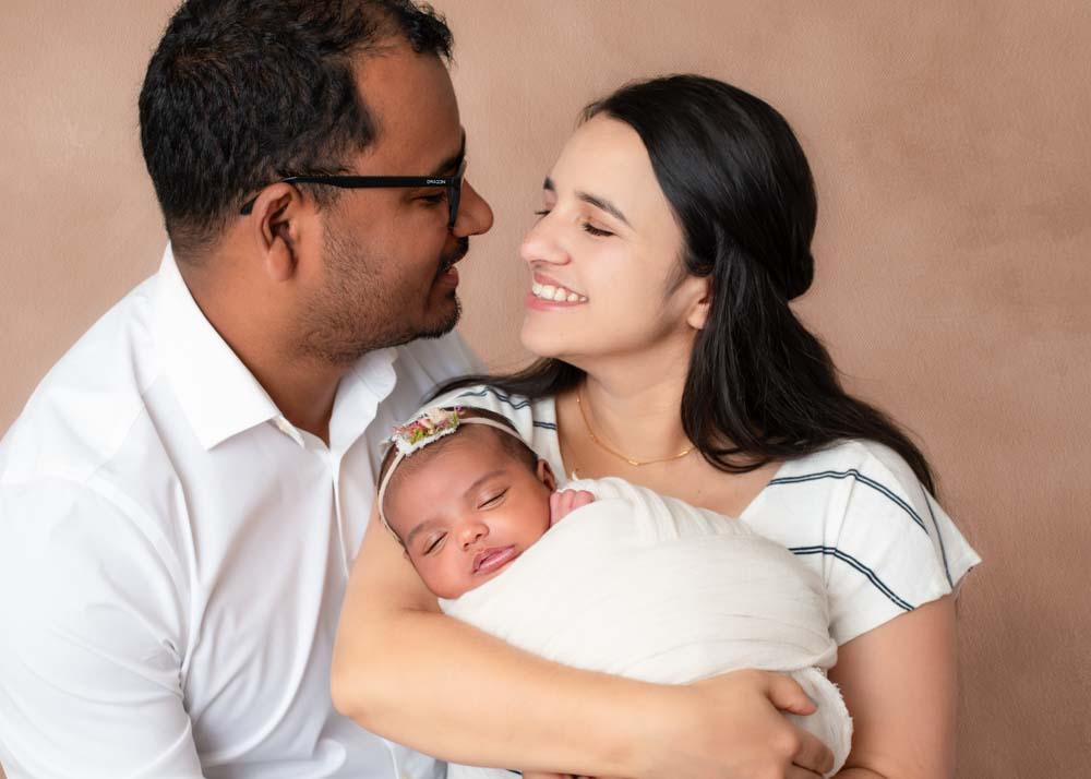 Parents smiling lovingly at each other while holding their swaddled newborn baby girl, posed against a soft beige studio backdrop in Birmingham newborn photography session