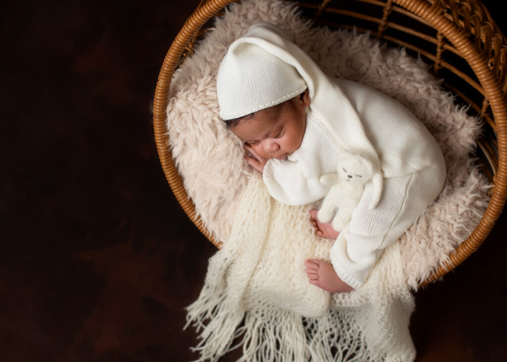 Newborn baby dressed in a white knitted outfit sleeping in a woven basket on a cream blanket — by Birmingham newborn photographer Misty Dawn Photography.