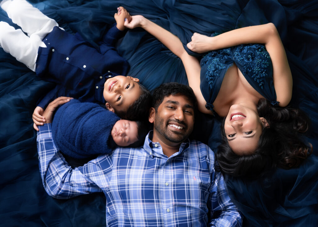 Newborn baby swaddled in a navy blue wrap lying next to their smiling older brother and parents on a dark blue backdrop — by Birmingham newborn photographer Misty Dawn Photography.