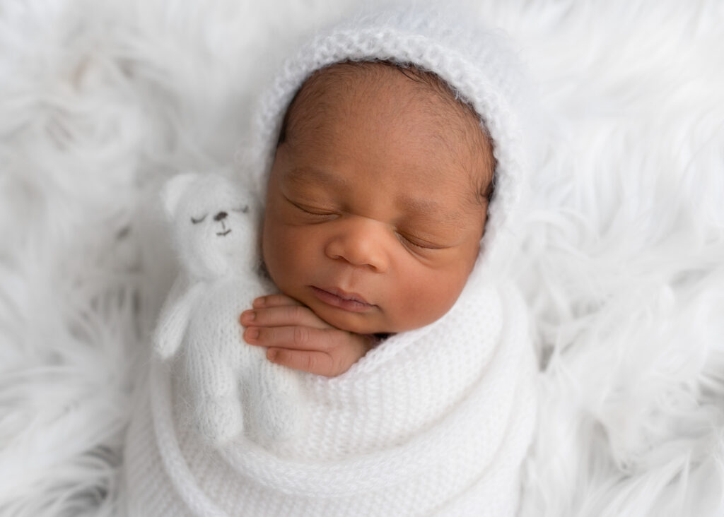 Newborn photo of baby wrapped in white knit and holding white teddy bear while resting on white fur.