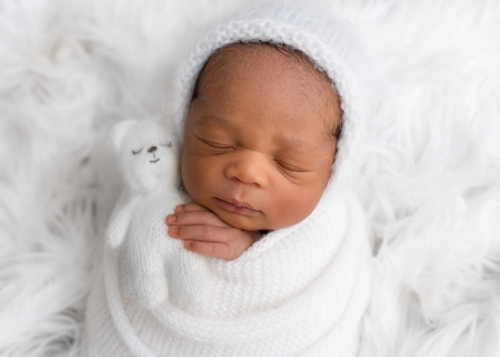 Newborn photo of baby wrapped in white knit and holding white teddy bear while resting on white fur.