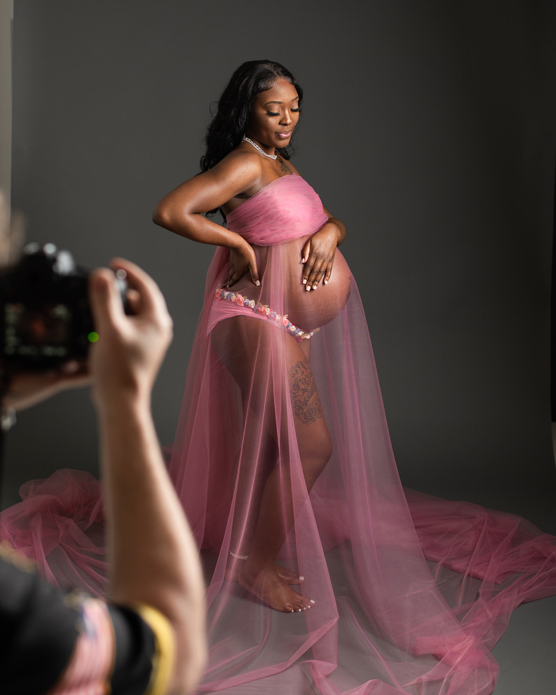 Photographer adjusting sheer fabric wrap during a flowing maternity pose in studio.