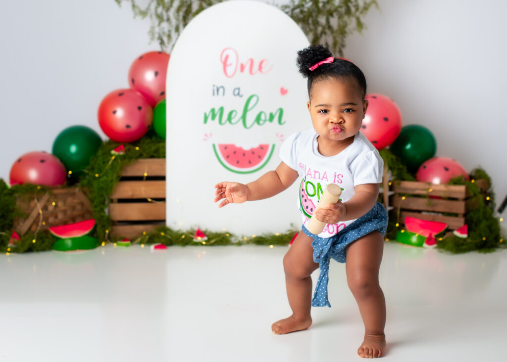 Baby girl standing and smiling in a watermelon-themed outfit during her One in a Melon cake smash session
