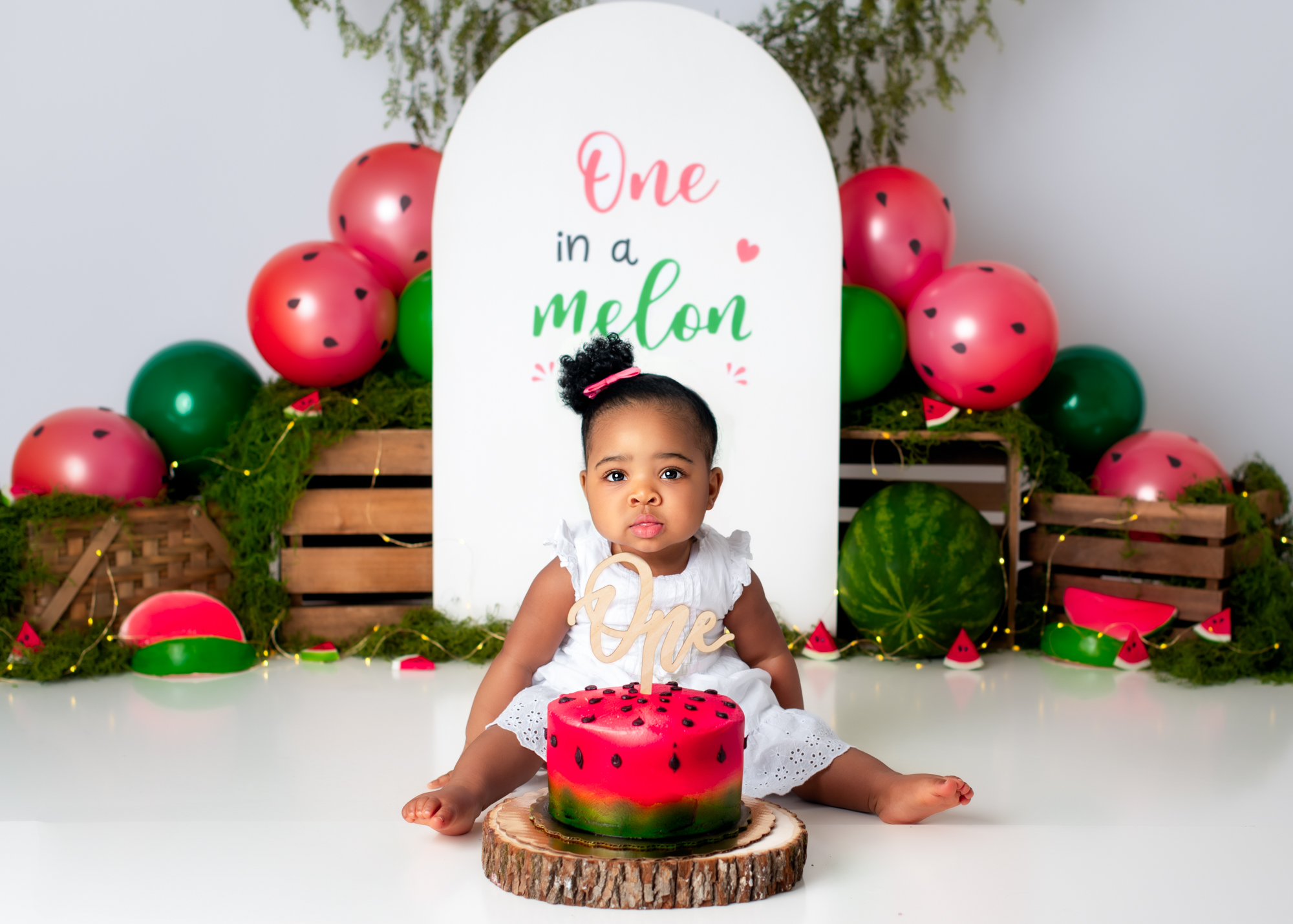 Baby girl sitting beside a red watermelon cake, backdrop with balloons and themed signage.