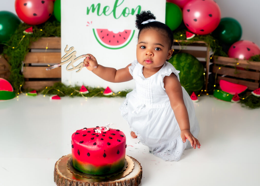 Baby girl in white dress examining her red cake during a watermelon-themed first birthday session.