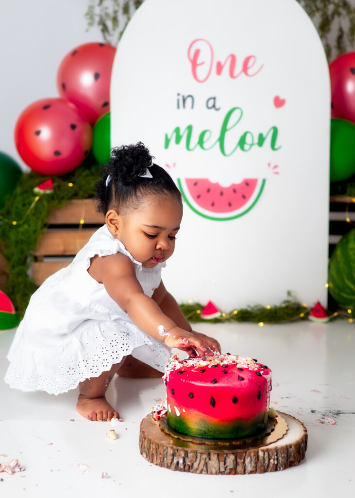 Baby in white dress playfully smashing watermelon-themed cake with both hands.