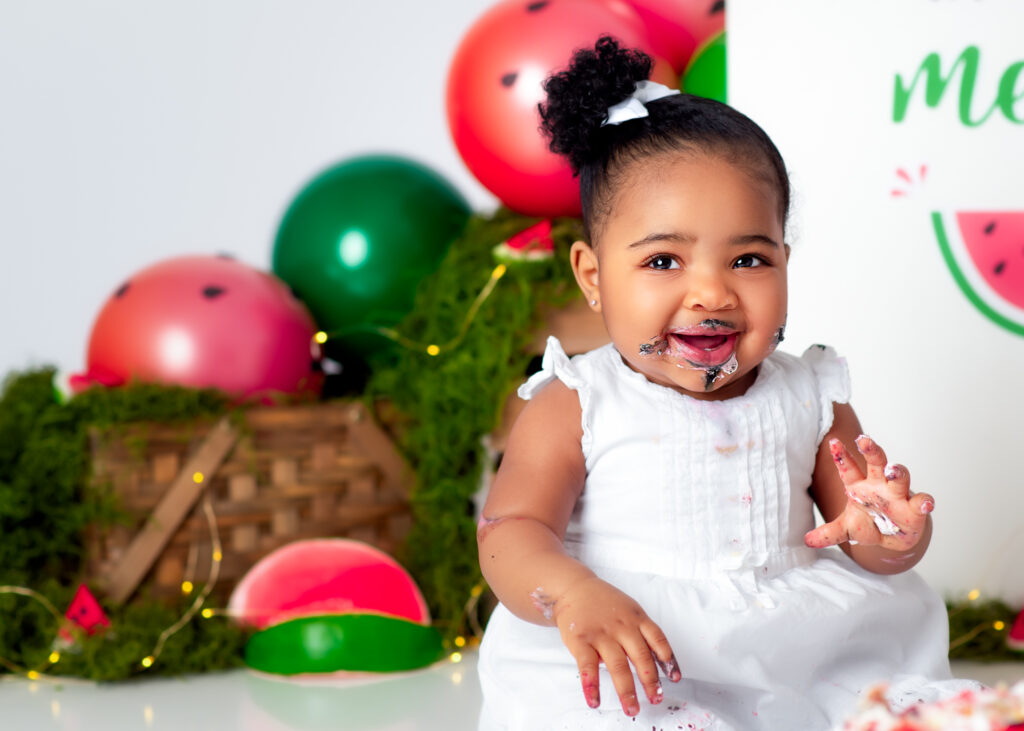 Close-up of baby with frosting on her lips, sitting in front of watermelon-themed birthday background.