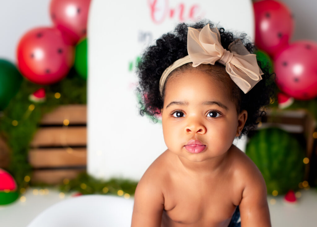 Close-up of toddler with curly hair and bow, seated in front of bright watermelon backdrop