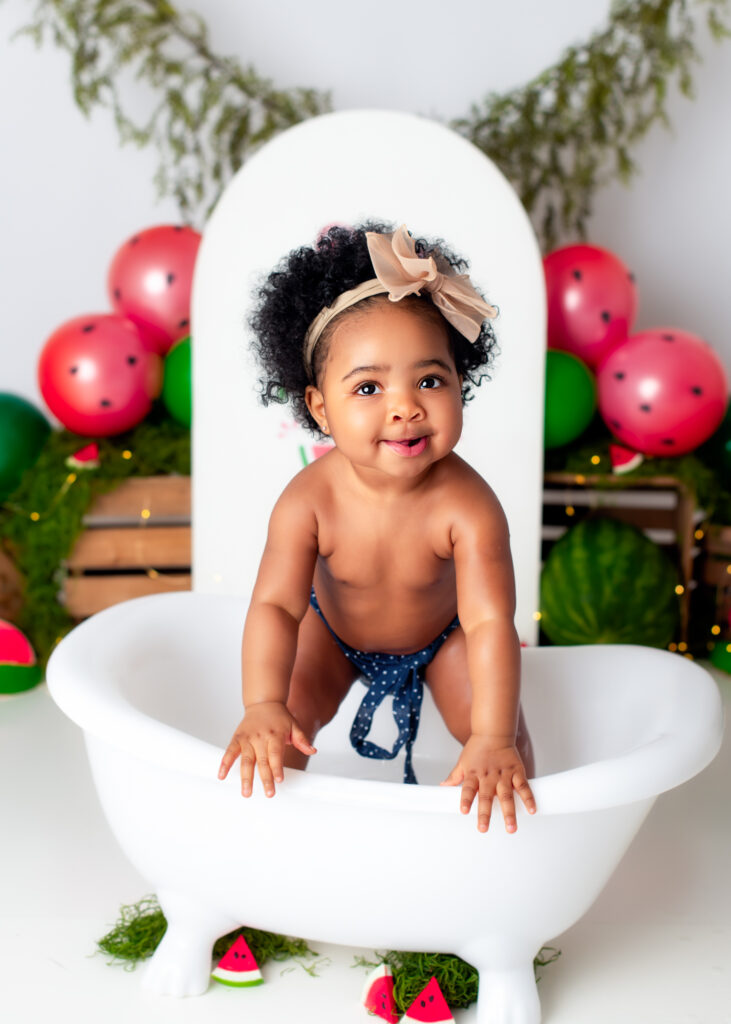 Smiling baby girl holding onto edge of white tub, looking up mid-splash in watermelon-themed setup