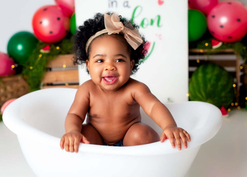 Toddler peeking over tub edge, smiling wide with bold summer decor and balloons behind her..