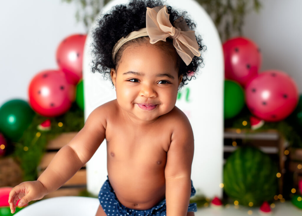 Baby girl smiling sweetly at camera, wearing a bow and blue diaper cover, red balloons in background.