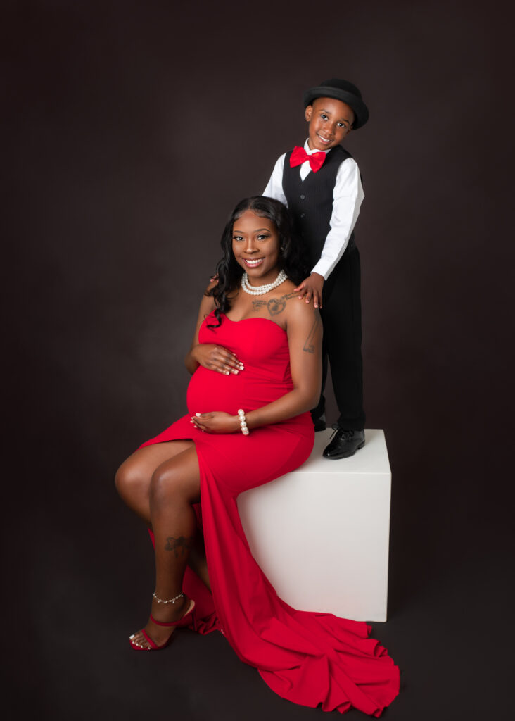 Studio maternity portraits featuring an expecting mother in a red gown with her partner and child, captured in elegant poses with dark seamless backdrop.