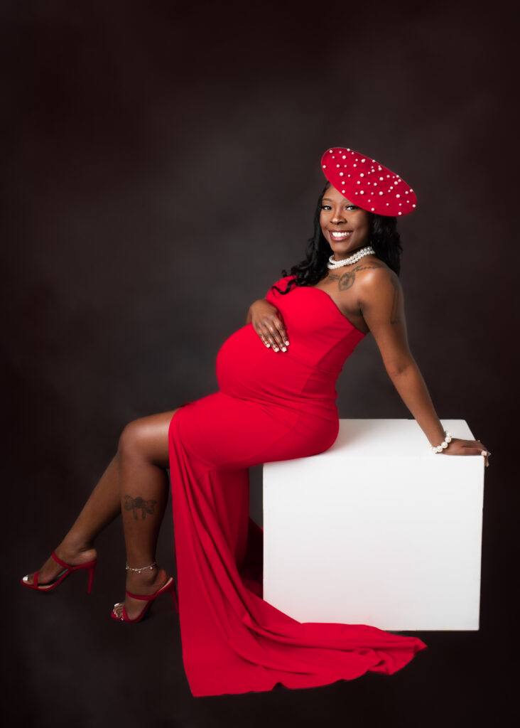 Maternity studio portraits of an expecting mom in a red gown and polka dot hat, styled for a bold, editorial look.