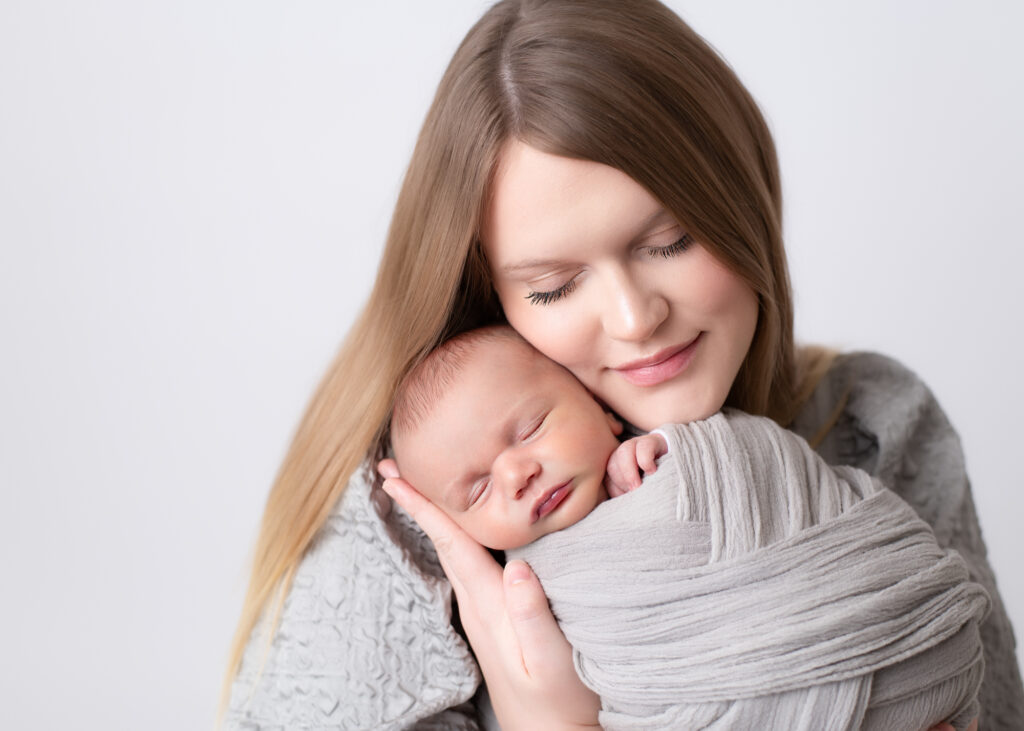 Mother holding her newborn baby close, both with eyes closed in peaceful connection during studio portrait session.