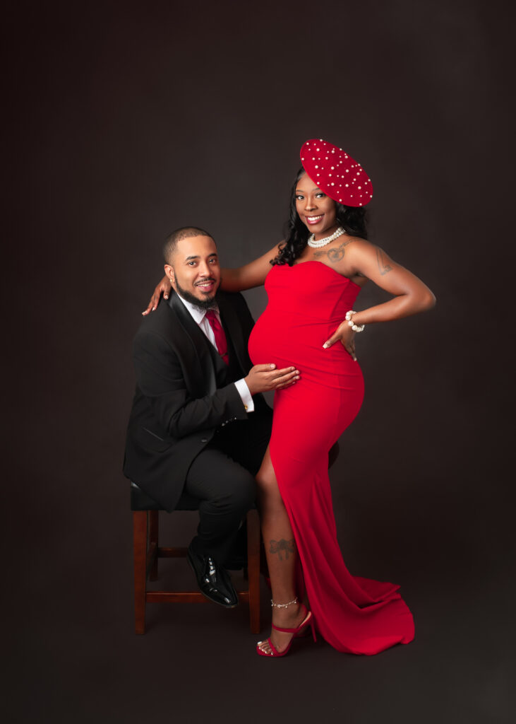 Studio maternity portraits featuring an expecting mother in a red gown with her partner and child, captured in elegant poses with dark seamless backdrop.