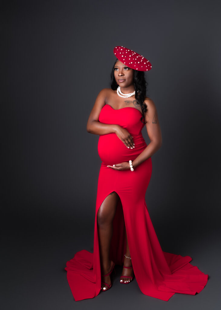 Maternity studio portraits of an expecting mom in a red gown and polka dot hat, styled for a bold, editorial look.