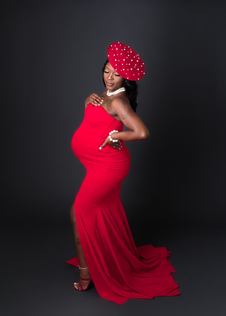 Maternity studio portraits of an expecting mom in a red gown and polka dot hat, styled for a bold, editorial look.