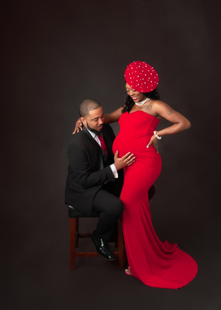 Studio maternity portraits featuring an expecting mother in a red gown with her partner and child, captured in elegant poses with dark seamless backdrop.