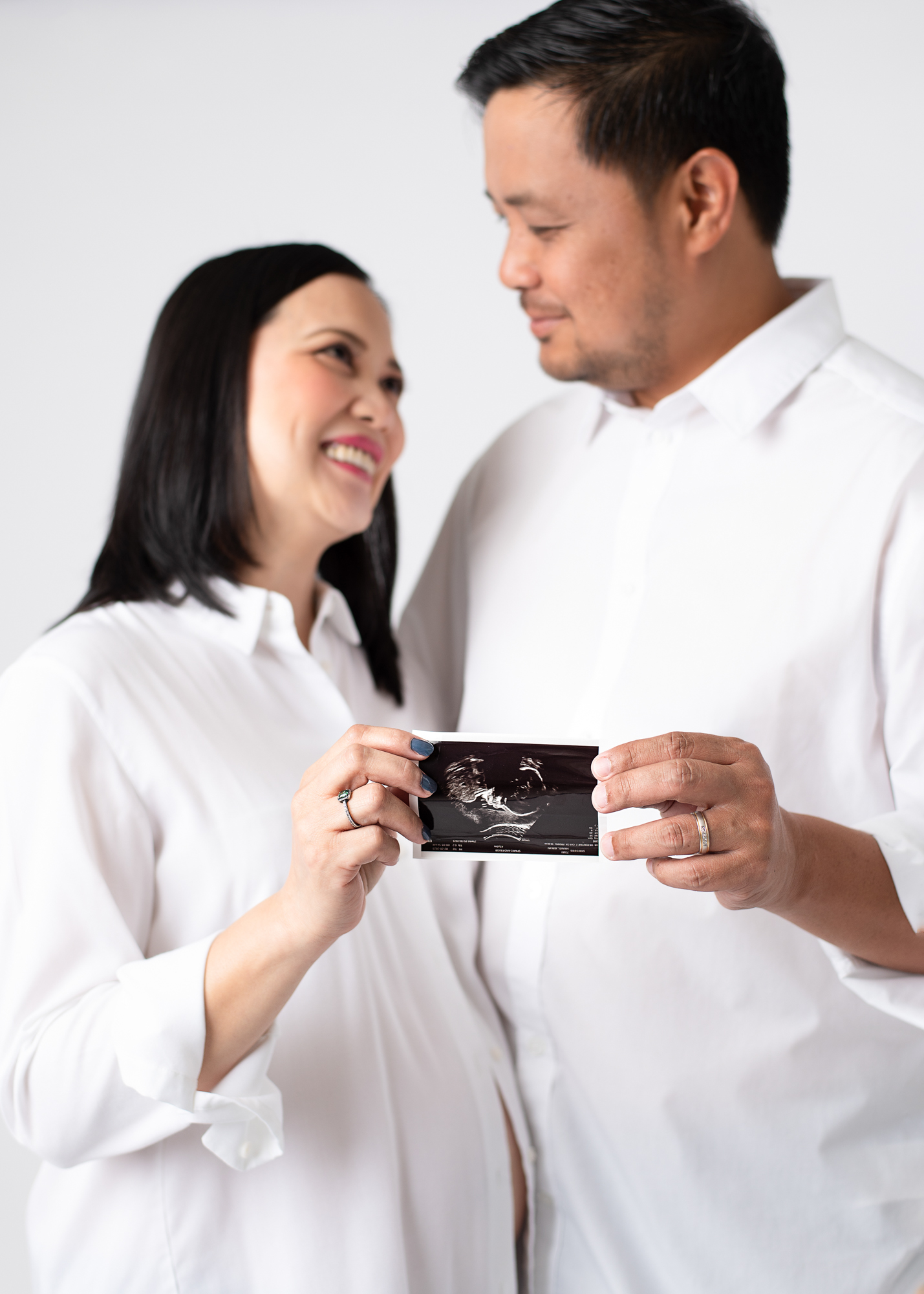 Expectant couple smiling at each other while holding a sonogram photo during a studio maternity session, dressed in coordinated white shirts.