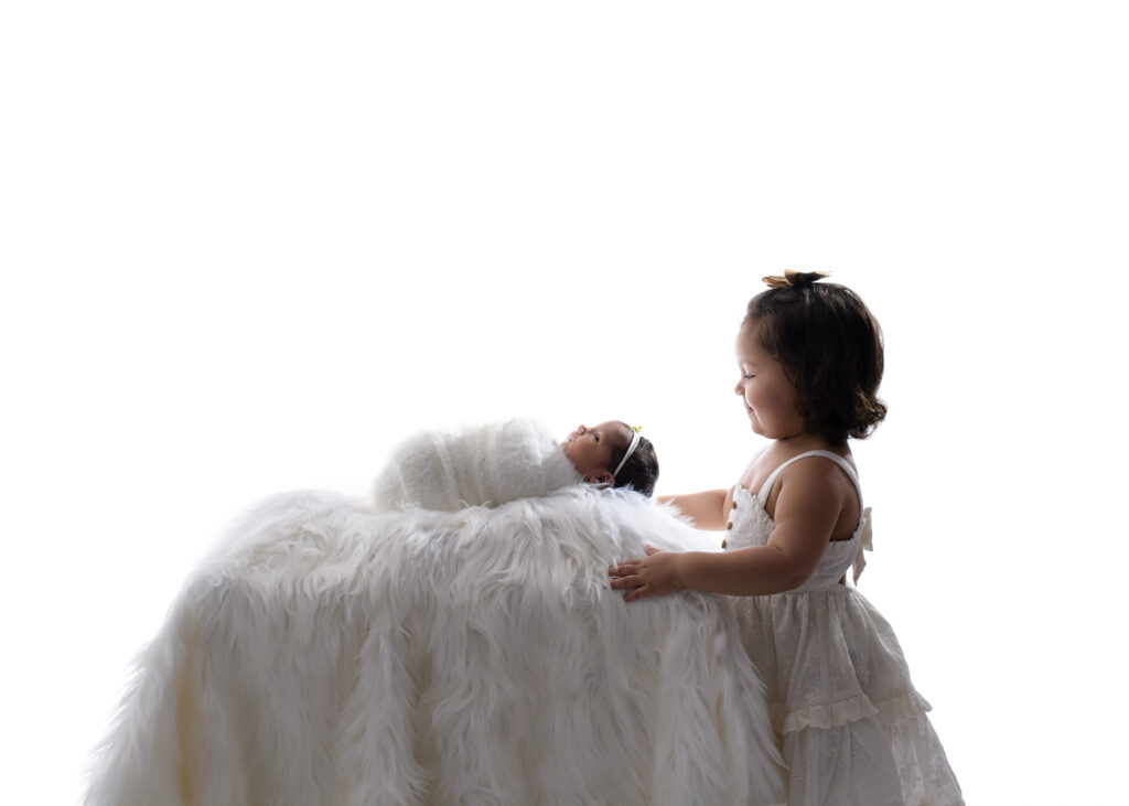 Toddler girl in a white dress smiling gently at her newborn sibling, who is swaddled and resting on a white faux fur blanket in a bright studio setting