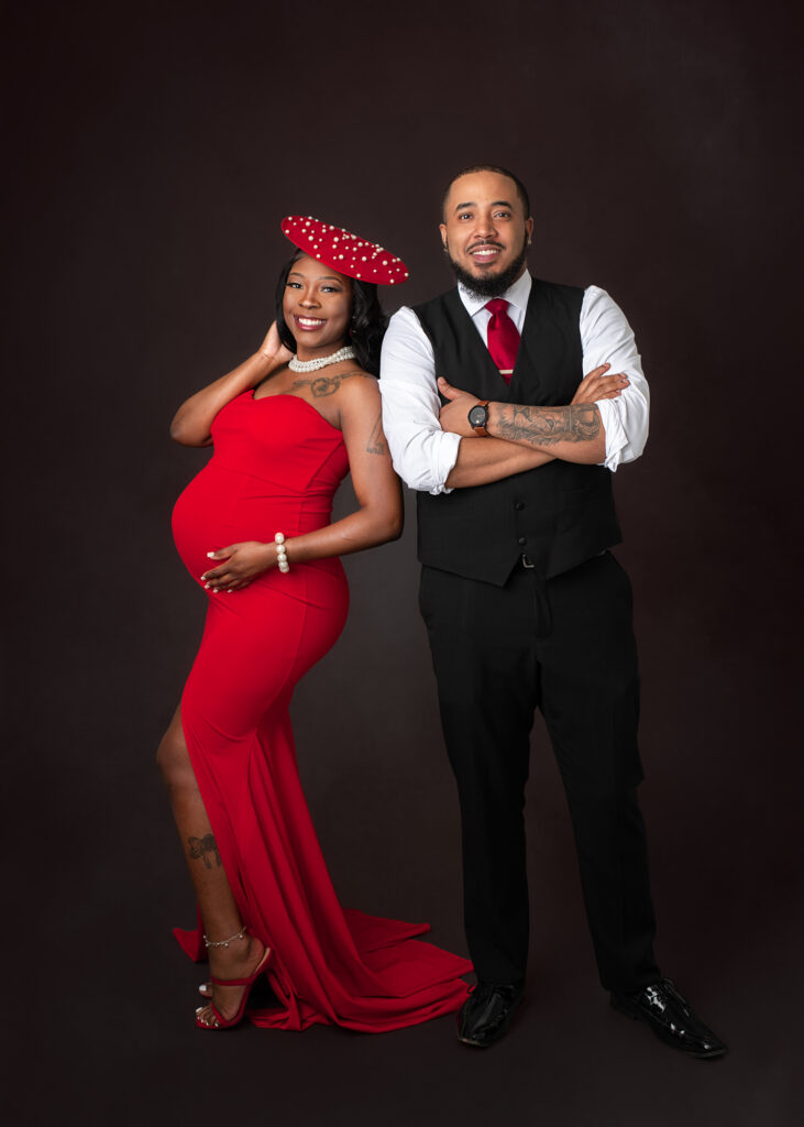 Studio maternity portraits featuring an expecting mother in a red gown with her partner and child, captured in elegant poses with dark seamless backdrop.