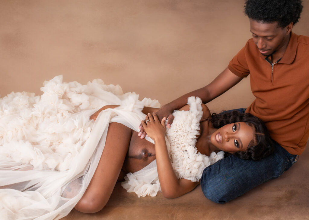 Studio maternity portrait of an expecting mother lying in a ruffled gown, embraced by her partner on a warm-toned backdrop.