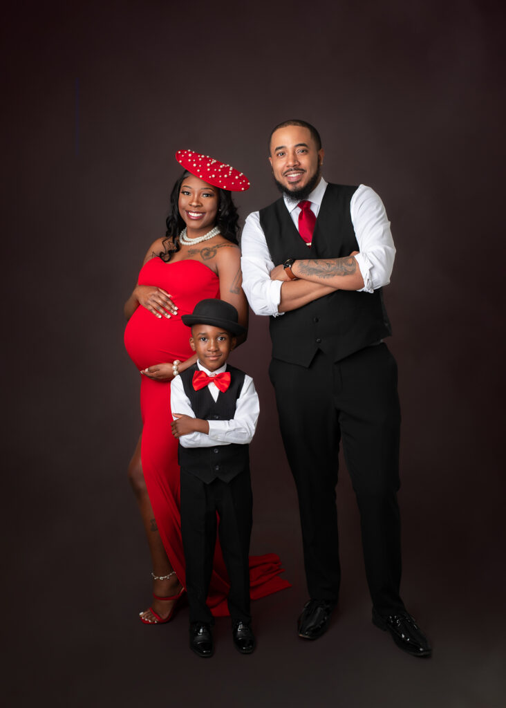 Studio maternity portraits featuring an expecting mother in a red gown with her partner and child, captured in elegant poses with dark seamless backdrop.
