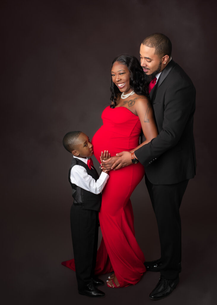 Studio maternity portraits featuring an expecting mother in a red gown with her partner and child, captured in elegant poses with dark seamless backdrop.