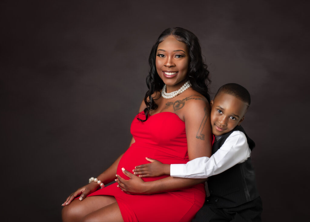 Studio maternity portraits featuring an expecting mother in a red gown with her partner and child, captured in elegant poses with dark seamless backdrop.