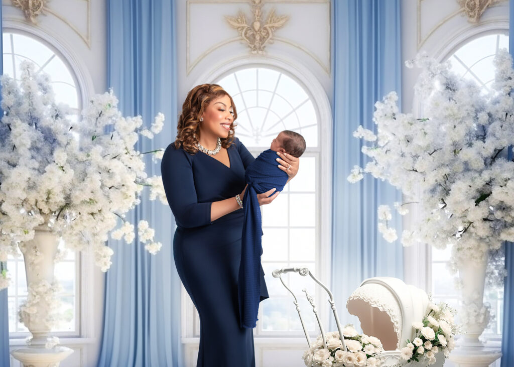 Studio portrait of a new mother in a navy gown holding her swaddled newborn, surrounded by white florals and soft natural light in a Birmingham photography studio.