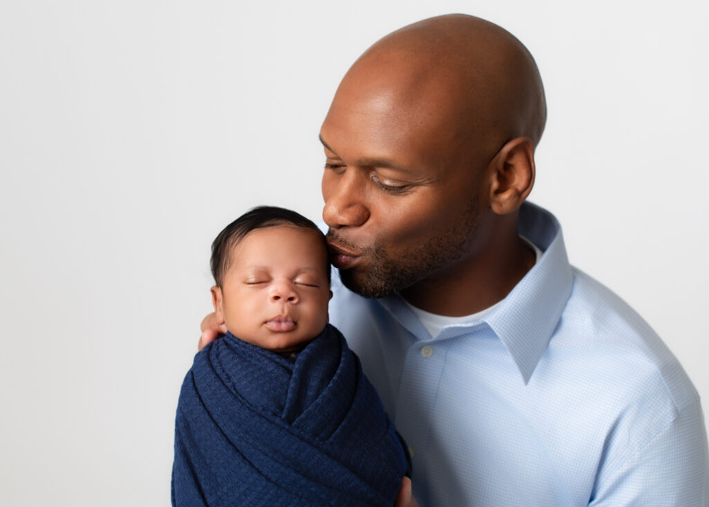 Studio portrait of a father kissing his baby’s forehead, swaddled in navy — captured by a newborn photographer in Birmingham.