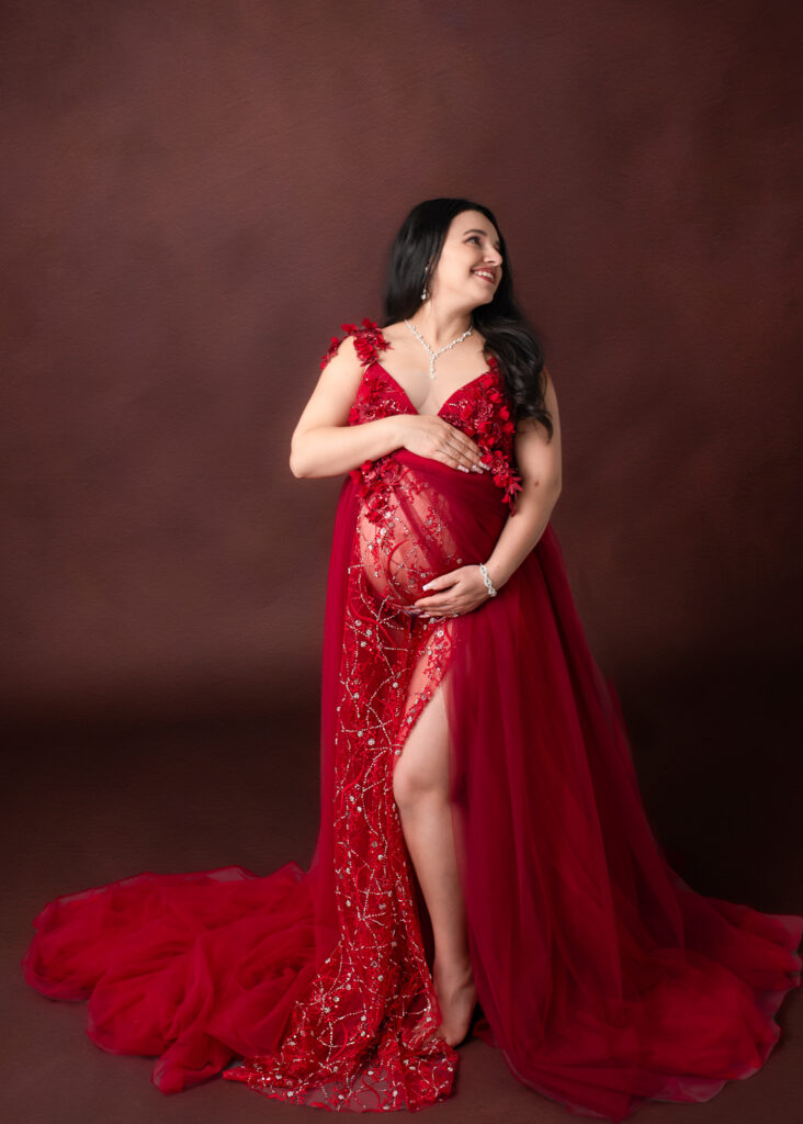 Studio maternity photo of a pregnant woman in a rich red gown with sparkling embellishments, standing against a deep brown backdrop — winter styling by a maternity photographer in Birmingham.