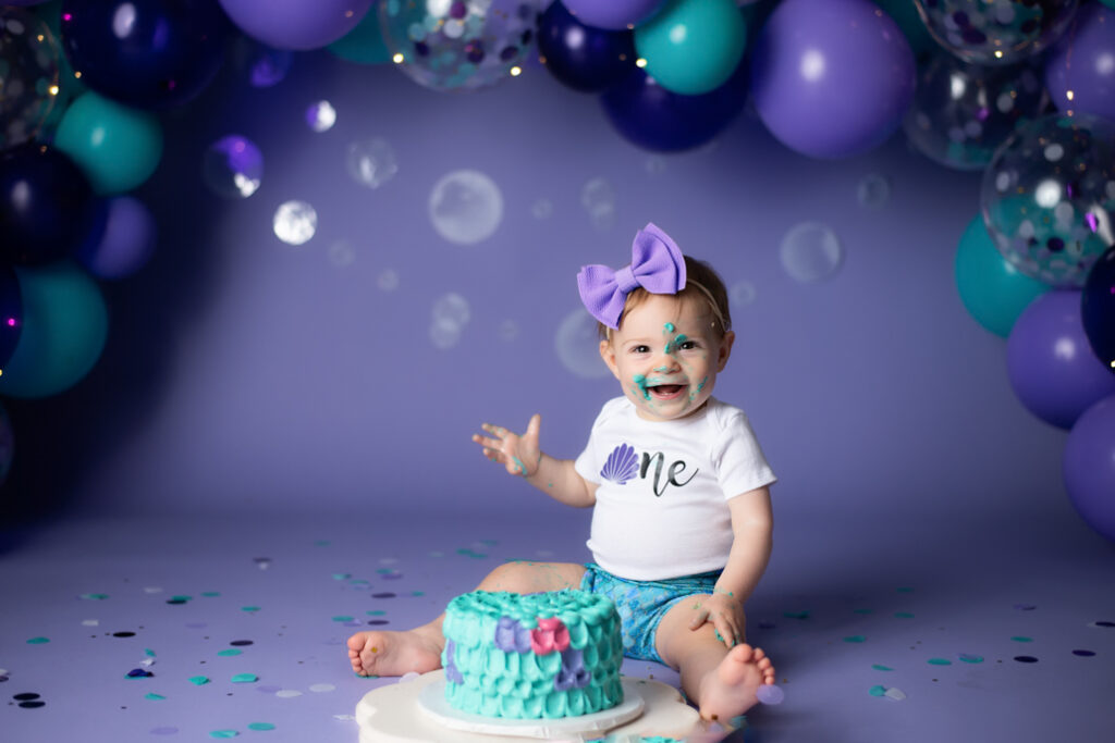 Mermaid-themed first birthday photoshoot with baby girl in “One” onesie and teal/purple confetti