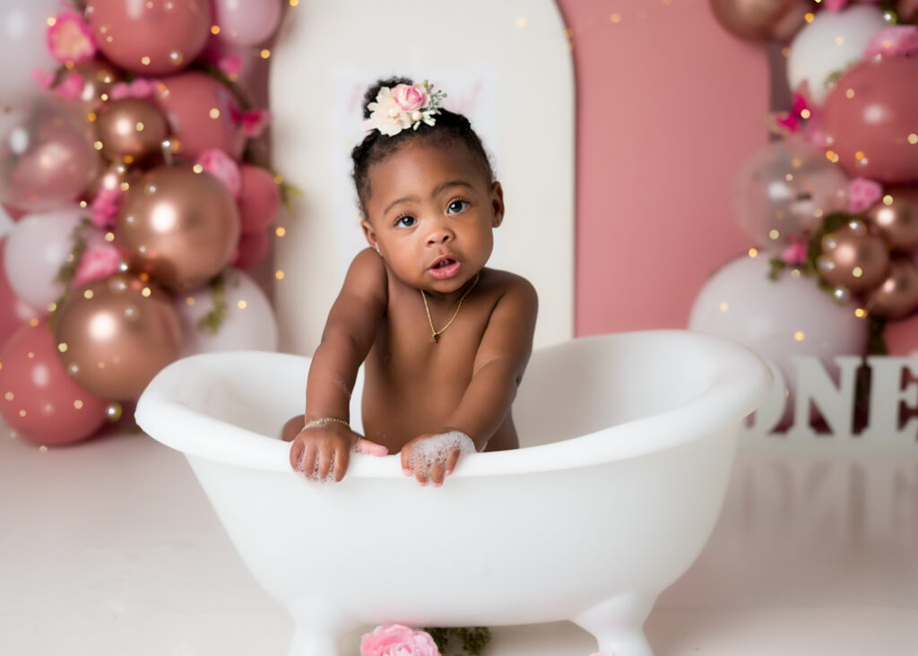 Little girl sitting in a white tub during splash session, smiling with bubbles and pink balloon garland behind