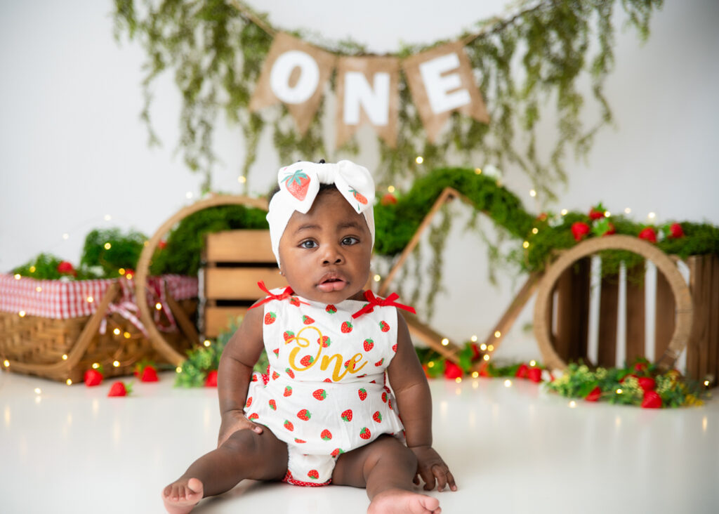 Baby girl in strawberry romper during strawberry-themed first birthday photoshoot with “ONE” sign and greenery