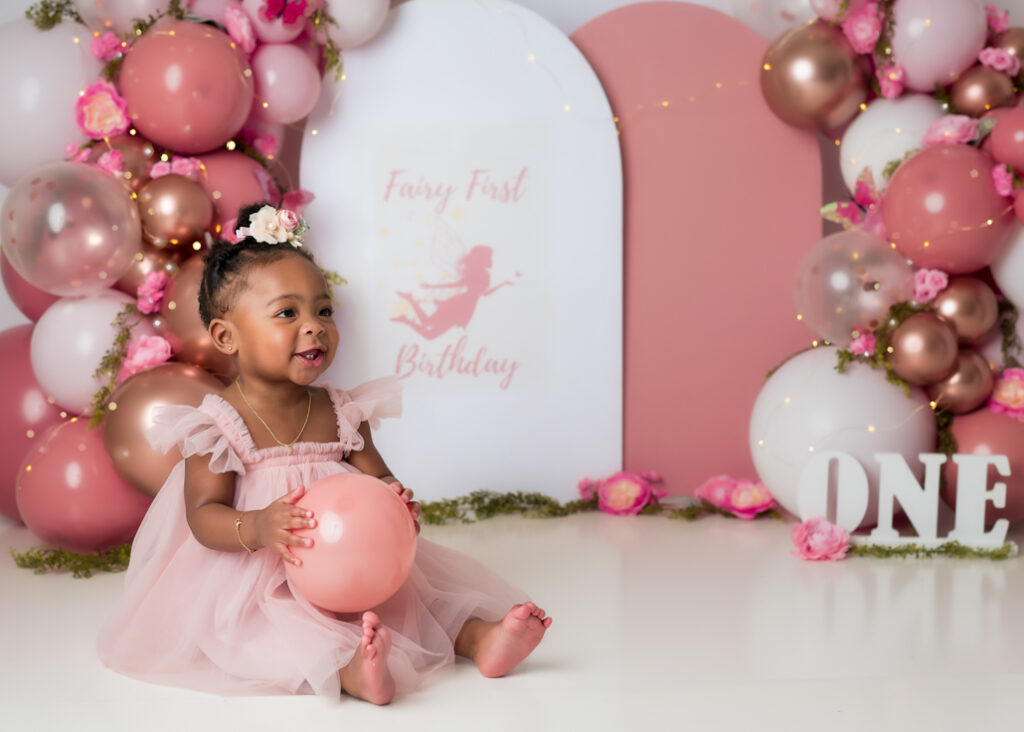 Smiling toddler in blush dress sitting with a balloon during a Fairy First Birthday photoshoot setup