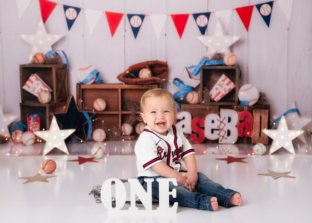 Smiling baby boy in Braves jersey sitting in front of baseball-themed setup with crates, bats, and red, white, and blue decorations