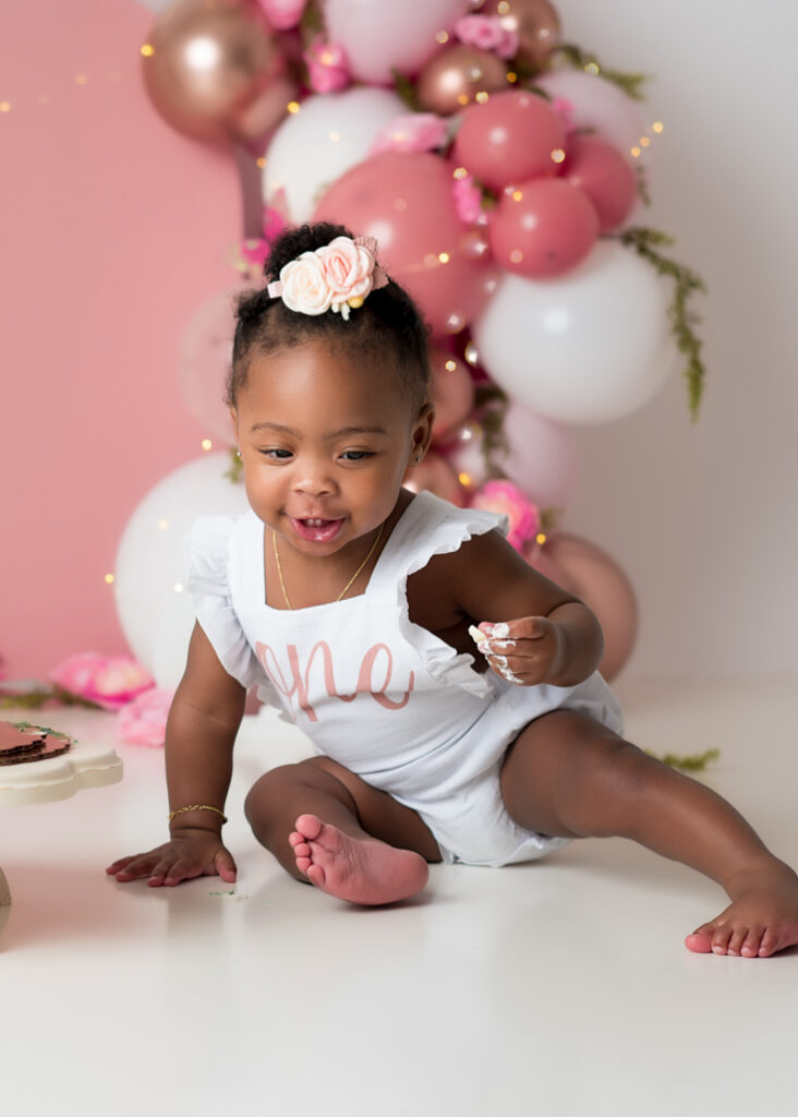 Toddler in white 'One' onesie smiling during cake smash session, sitting in front of fairy birthday setup