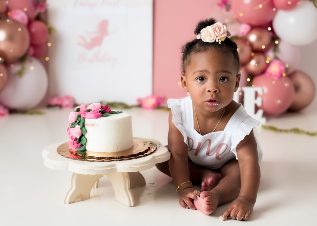 Baby girl seated next to white cake on a stand, ready for her cake smash with soft pink and floral decorations