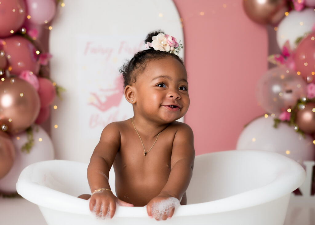 Toddler looking up with joy during splash portion of first birthday session, seated in mini white bathtub